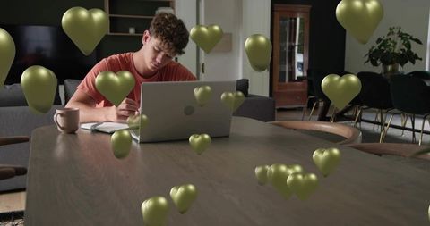 Young man working from home on laptop surrounded by floating gold heart balloons