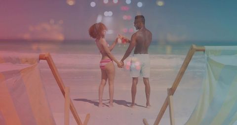 Couple toasting cocktails on tropical beach while holding hands by deck chairs with sunlit horizon