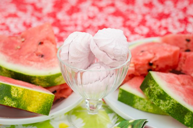 Sweet marshmallows displayed in a glass cup with fresh watermelon slices on a vibrant, colorful table. Perfect for representing summer desserts, refreshing treats, and seasonal snacks. Ideal for food blogs, recipe sites, or summer party promotions.