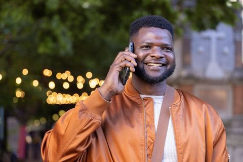 Smiling african man talking on phone in city evening with bokeh lights and bomber jacket
