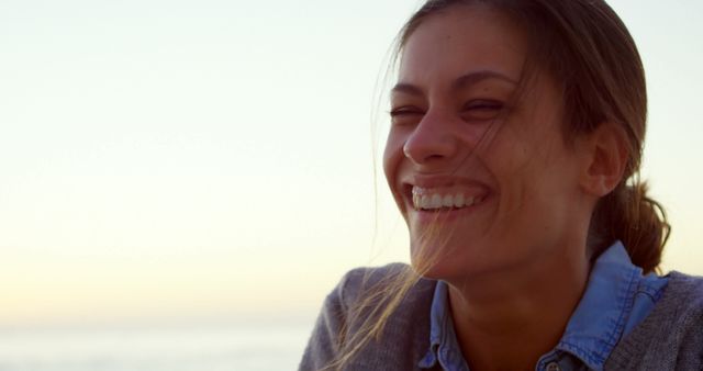 Young woman smiling joyfully by the seaside during sunset. Perfect for themes of happiness, relaxation, outdoor activities, and positive emotions. Ideal for use in advertisements, travel brochures, wellness programs, and lifestyle blogs.