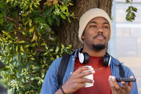 African american man standing by tree holding coffee and phone wearing beanie and headphones