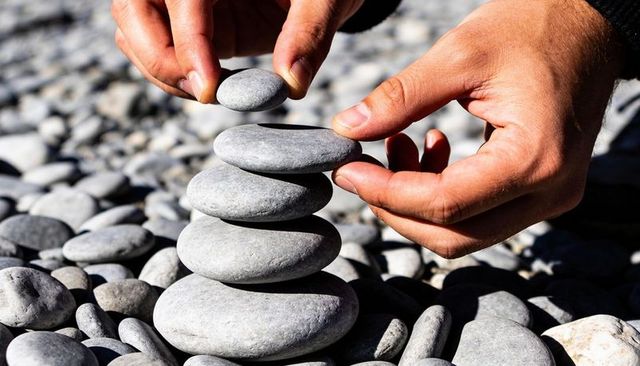 Placing and balancing pebble cairn, male hands stacking five smooth stones on sunlit pebble beach
