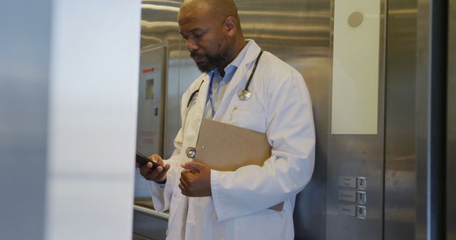 This image shows an African American doctor wearing a white coat and using a smartphone while standing in an elevator. He holds a clipboard, indicating that he is engaged in professional tasks. Perfect for use in medical or healthcare-related articles, advertisements for medical services, or brochures for hospital facilities to emphasize modern and up-to-date healthcare practices.