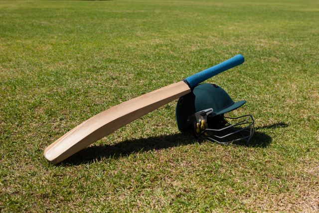 Cricket bat and helmet lying on grass field during a sunny day. Ideal for use in sports-related content, cricket match promotions, safety gear advertisements, and outdoor recreation themes.