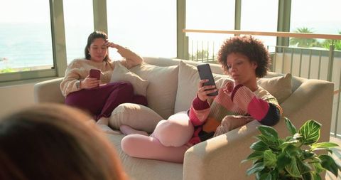 Multiracial women lounging and scrolling smartphones on sunlit coastal sofa with ocean view