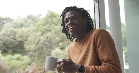 African american man smiling by window holding coffee mug in cozy sweater morning light