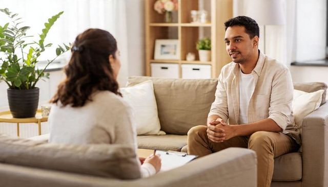 Male client attending therapy session in cozy living room, counselor taking notes