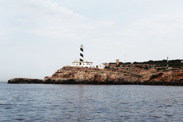Lighthouse standing on a rocky coastline, bordered by calm blue waters and an expansive sky. Ideal for themes related to travel, tourism, maritime navigation, coastal living, and serene landscapes. Perfect for travel brochures, website headers, or inspirational posters.