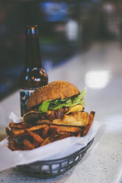 This image depicts a close-up of a gourmet burger with fries and a bottle of beer in a restaurant setting. Ideal for use in marketing materials for restaurants, ads for dining establishments, food blogs, or social media posts promoting upscale fast food.