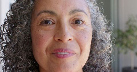 Gazing senior woman with curly gray hair smiling and showing freckles by sunlit door