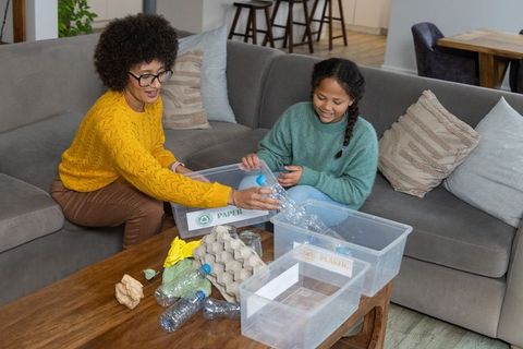 Mother and daughter sorting recycling together on couch with labeled plastic and paper bins