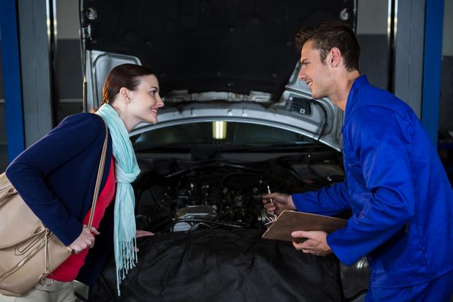Mechanic in blue uniform explaining repair quotation to female customer at a car repair garage. Customer leaning forward, showing interest in the explanation. Useful for illustrating automotive services, customer service interactions, and professional consultations in repair shops.