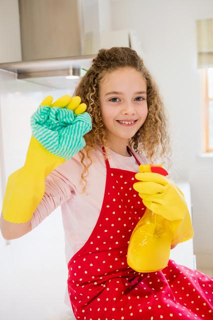 Young girl with curly hair smiling while holding a spray bottle and napkin in a kitchen. She is wearing yellow gloves and a red polka dot apron, indicating she is engaged in household chores. This image can be used for promoting cleaning products, household chores, family activities, and hygiene practices.