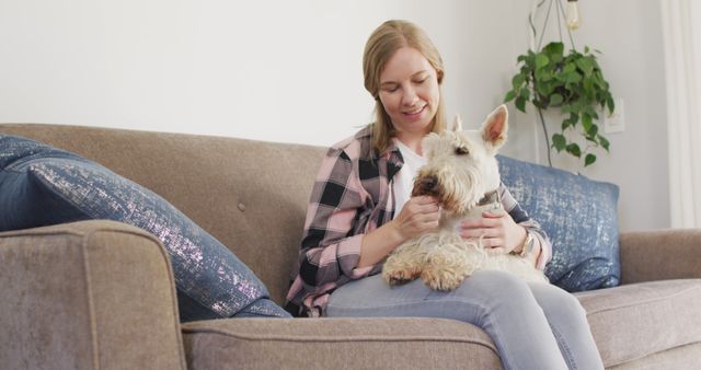 A young woman is sitting on a comfortable couch in a modern living room, happily petting her Westie dog. This relaxed and homely scene showcases companionship and bonding between her and her pet. Perfect for use in advertisements related to pet care, home decor, cozy lifestyles, or blog posts about pets and comfort at home.