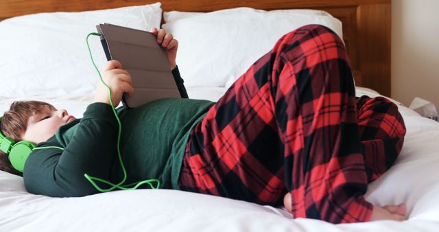 Young boy lying on bed wearing red plaid pajamas pants and green shirt, listening to music with green headphones while using a tablet. Perfect for technology related content, child leisure activities, home life blogs, and advertisements focused on digital devices for kids.