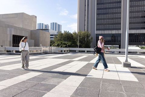 Two women walking across sunlit urban plaza with backpacks, coffee and smartphones