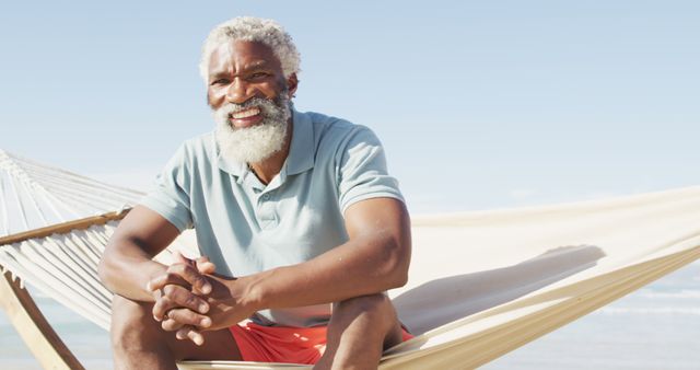 Portrait of happy senior african american man sitting in hammock on sunny beach. healthy and active time beach holiday.