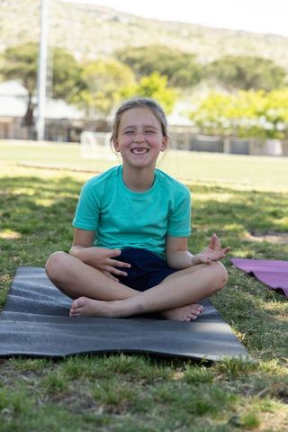 Young girl sitting cross-legged on yoga mat barefoot smiling outdoors in sunny park field