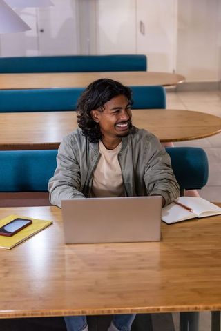 Young man smiling while working on laptop with notebook and phone in modern coworking cafeteria