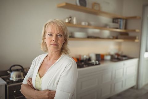 Senior woman stands with arms crossed, expressing confidence in a modern kitchen environment. Elegant accents like wooden shelves and a silver kettle contribute to a cozy and minimalist aesthetic. Ideal for use in advertisements or brochures promoting lifestyle tips, senior living, or homemaking advice centered around elegance and comfort.