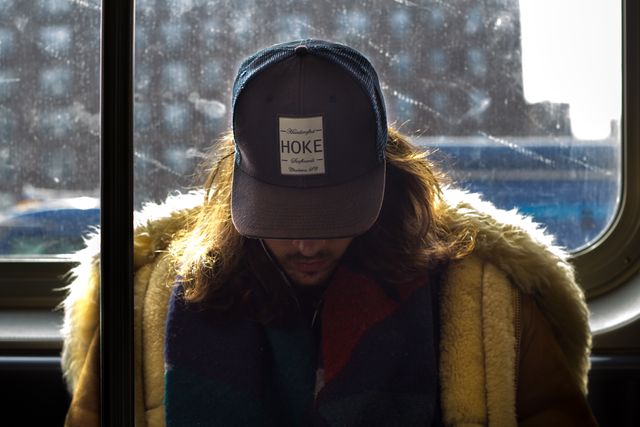 The young man is wearing a hat and warm winter coat while sitting on a bus. His face is obscured by the brim of the hat and he seems lost in thought while looking downward. The background shows the city through the bus window with reflections suggesting movement. It is a serene moment in daily urban commuting. Suitable for use in articles about city life, public transportation, youthful fashion, or introspective moments in urban settings.