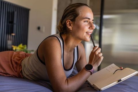 Young woman journaling on bed, contemplating ideas with pen and smartwatch in daylight