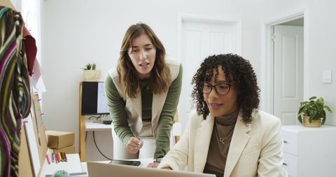 Mid-adult female coworkers collaborating over laptop in bright modern office, pointing, planning