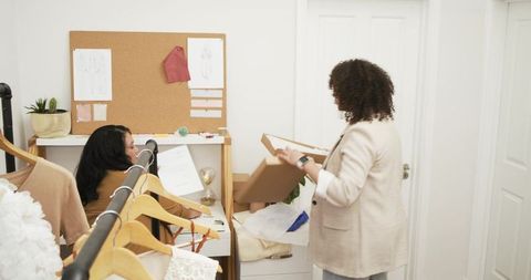 Diverse female coworkers packing garments and reviewing sketches in boutique backroom