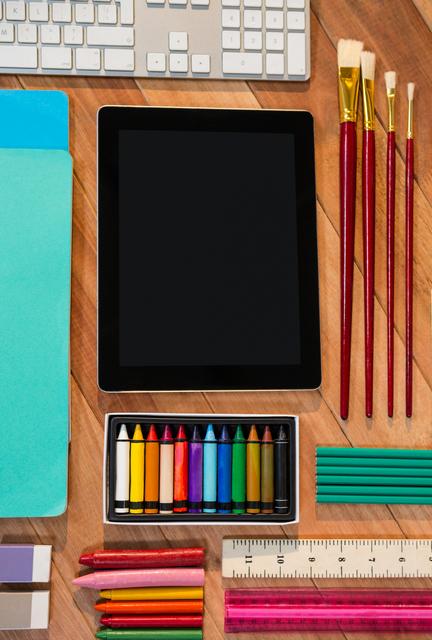 Digital tablet surrounded by various stationery items including crayons, paintbrushes, keyboard, and rulers on a wooden table. Ideal for use in educational content, back-to-school promotions, creative workspace setups, and technology integration in classrooms.