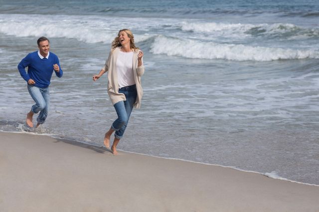 Mature couple running and laughing on sandy beach near ocean waves. Ideal for promoting active lifestyles, vacation destinations, relationship bonding, and outdoor leisure activities.