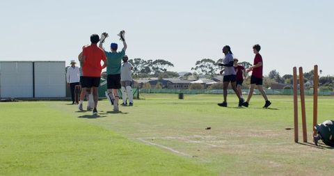 Local cricket team celebrating wicket on sunny outfield with umpire, stumps and helmets