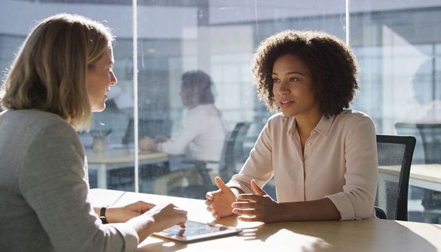 Businesswomen engaging in collaborative conversation at sunlit glass office table