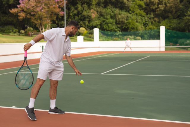 Two Caucasian men wearing tennis whites spending time on a court playing tennis on a sunny day, holding a tennis rackets, one man bouncing ball. Hobby sport leisure time.