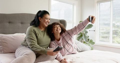 Two Women Laughing and Taking Selfie on Bed in Bright Cozy Bedroom