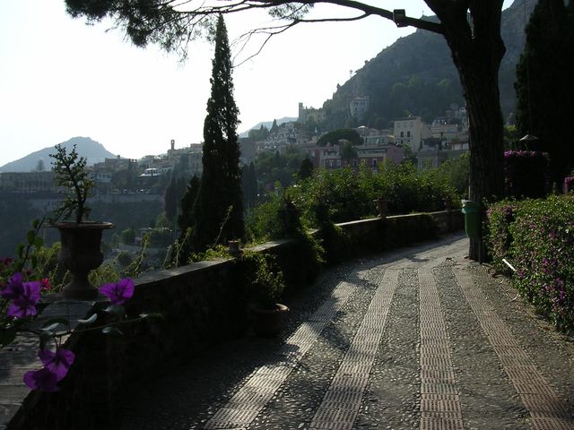 This image shows a picturesque walkway along a serene mountain village. The path is bordered by flowering trees and lush greenery. The village has a quaint, peaceful ambiance, making it an ideal setting for articles or promotions related to travel, relaxation, nature walks, and scenic destinations.
