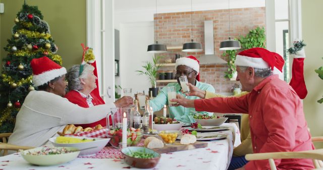 Seniors wearing Santa hats enjoying a festive meal together, clinking glasses and smiling at dining table with Christmas tree in background. Ideal for promoting holiday spirit, family gatherings, or festive season themes for cards, advertisements, and social media posts.