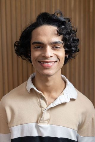 Teenage indian male smiling headshot beige polo hoop earrings warm wood slat background