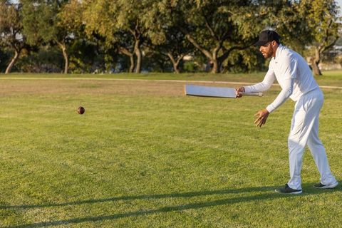 Cricketer practicing horizontal bat hold while ball is arcing over sunlit grass field