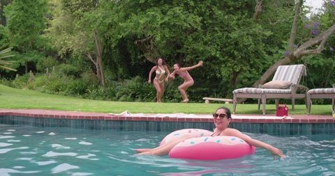 Woman relaxing on pink donut float in backyard pool with friends and lounge chairs, summer