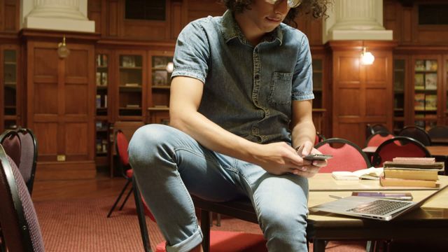 Young man casually sitting on table in library, smiling while using phone and laptop. Features wooden bookshelves and red chairs. Could be used to depict studying, education technology, digital learning, or relaxing moments in academic environment.