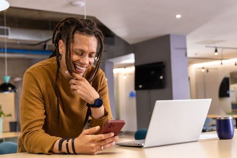 Smiling African American man leaning using smartphone beside laptop in modern coworking