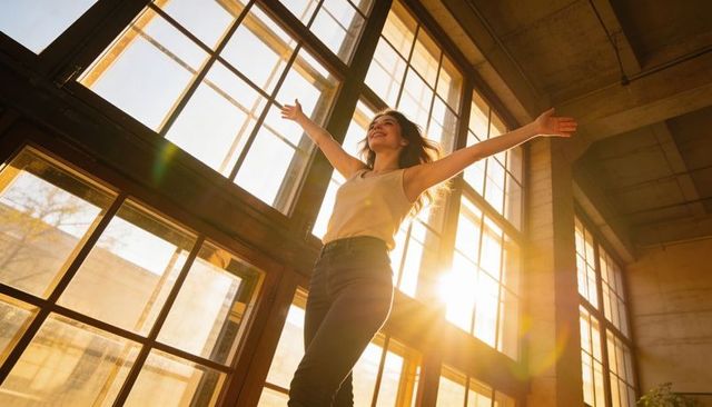 Smiling woman stretching arms in sunlit industrial loft with golden lens flare