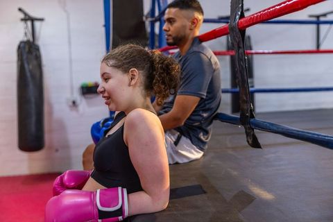 Teenage female boxer resting against ring ropes, trainer sitting beside, post-sparring recovery