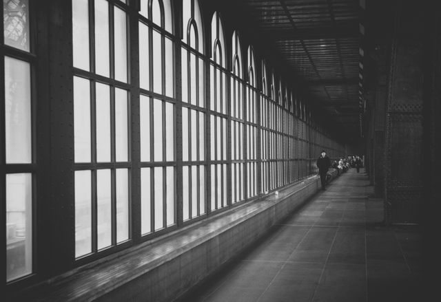 Depicting a long industrial walkway from a monochrome perspective, showcasing the contrast of shadows and lighting with large windows on one side and a person in the distance, highlighting the scale and symmetry. Useful for architecture, history, and urban-themed content, as well as concepts related to perspective and industrial aesthetics.