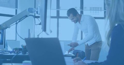 Bearded man leaning over laptop sharing phone in bright open-plan coworking office
