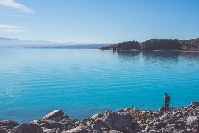 Hiker exploring rocky shores of a beautiful blue lake under a clear sky. Perfect for promoting outdoor activities, travel destinations, nature adventures, and serene landscapes. Ideal for use in blogs, websites, travel brochures, and social media posts.