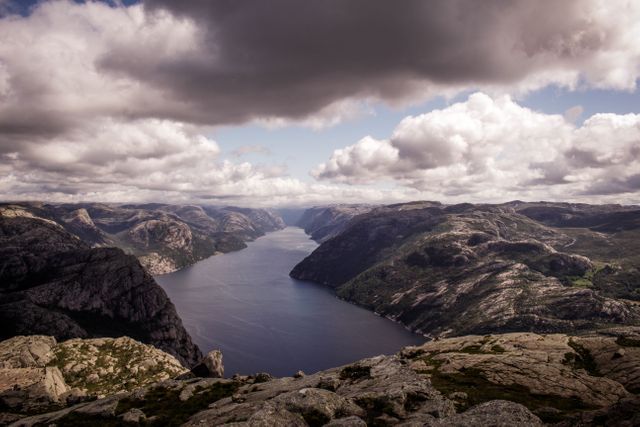 Panoramic view of a dramatic fjord with a majestic cloudy sky and rugged mountains. Ideal for travel brochures, nature magazines, and websites promoting adventure tourism. Captures the tranquility and grandeur of Norway's wilderness, perfect for outdoor adventure campaigns and scenic backgrounds for presentations.
