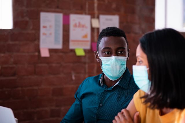 This image shows a diverse male and female colleague wearing face masks while discussing work at a desk in a modern office. It is ideal for use in articles or presentations about workplace safety, business operations during the COVID-19 pandemic, teamwork, and health protocols in professional settings.