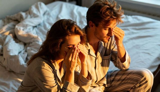 Couple sitting in pajamas rubbing eyes on unmade bed at sunrise, morning fatigue and intimacy